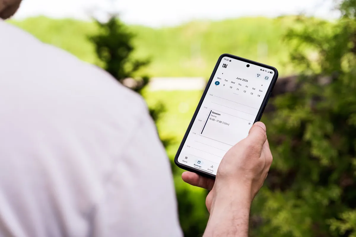 A person using a sauna booking app on a smartphone outdoors, surrounded by greenery.