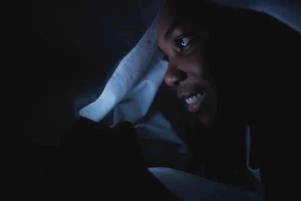 A woman smiles while using her smartphone under the covers in a dark room.