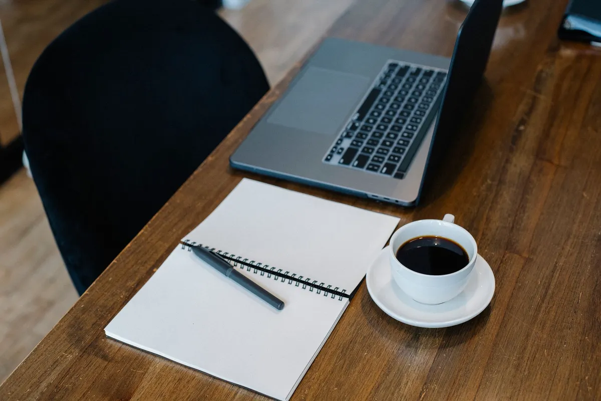 High angle of opened laptop and spiral notebook with blank pages placed on wooden table with cup of coffee and pen during workday
